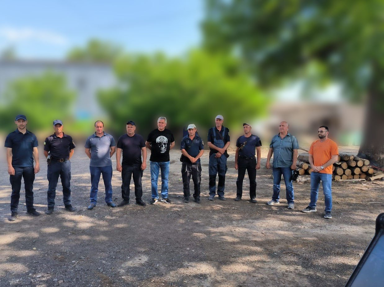Шеренга з десяти чоловіків стоїть на відкритому майданчику на тлі розмитих дерев та складених дров. Частина чоловіків ліворуч одягнена у формений одяг і кепки, інші — у цивільний одяг, як-от футболки, сорочки та джинси. Подія відбувається на вулиці у сонячний день.