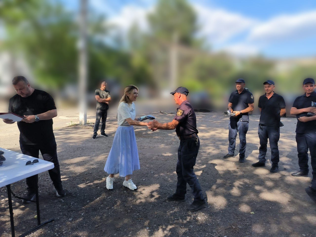 Жінка у білій футболці та довгій світло-блакитній спідниці тисне руку невисокому засмаглому чоловікові міцної статури у темній формі та кепці, вручаючи йому пакунок. Ліворуч біля столу стоїть чоловік із документами в руках, а праворуч у шерензі спостерігають інші колеги у формі. Подія відбувається на вулиці з розмитим фоном.