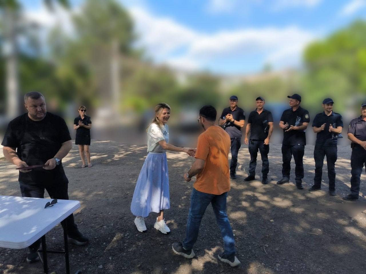 Жінка у білій футболці та довгій світло-блакитній спідниці посміхається і тисне руку чоловікові у помаранчевій футболці та джинсах. Ліворуч біля столу стоїть чоловік у чорному одязі, а праворуч у шерензі стоять та аплодують кілька осіб у темній формі. Захід проходить на вулиці у сонячний день на розмитому фоні зелені.