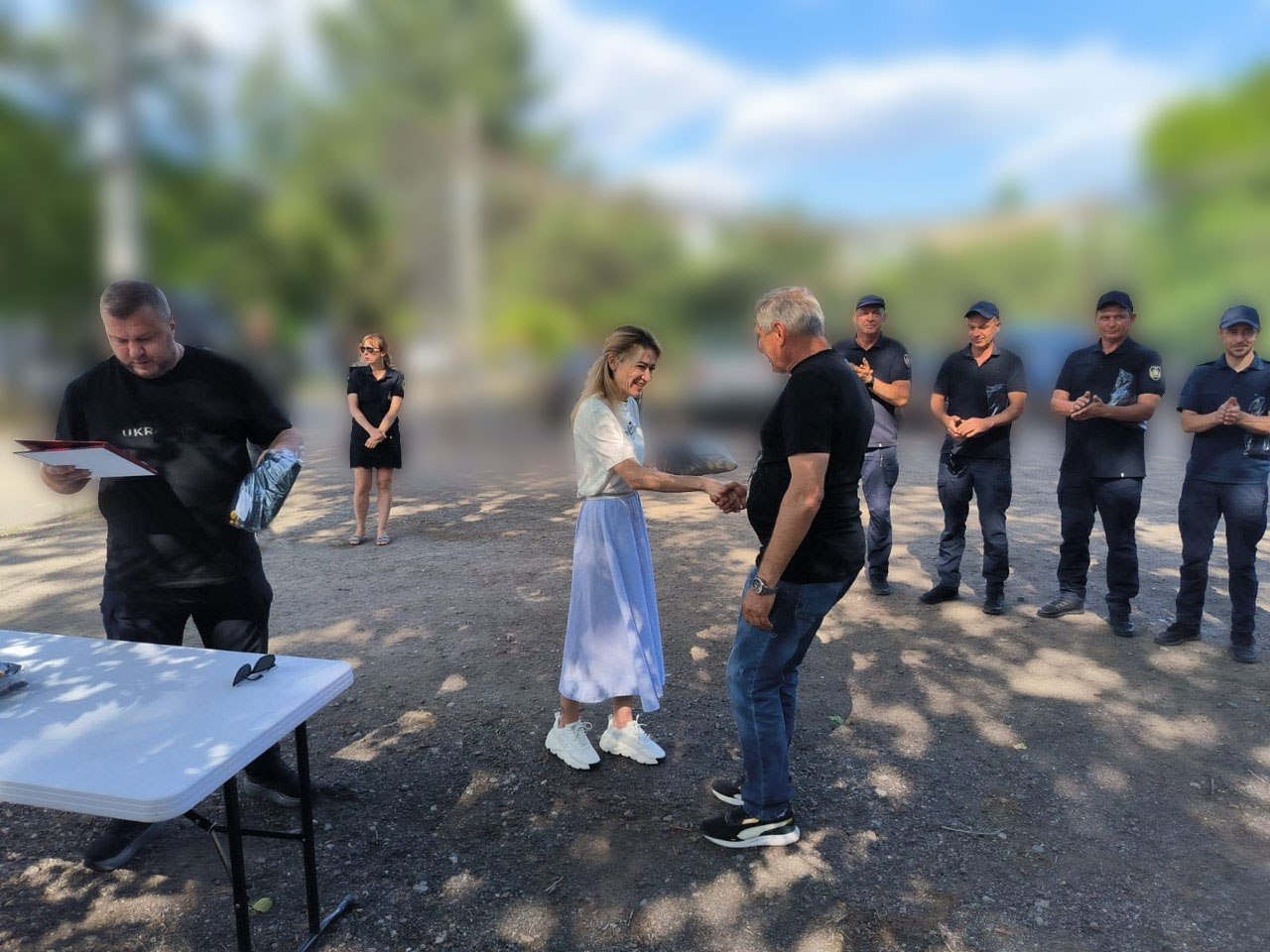 Жінка у білій футболці та довгій світло-блакитній спідниці тисне руку чоловікові у чорній футболці та джинсах на відкритому майданчику. Ліворуч стоїть чоловік біля столу і переглядає документи, а праворуч у шерензі стоять та аплодують четверо осіб у темній формі. Задній план зображення розмитий.