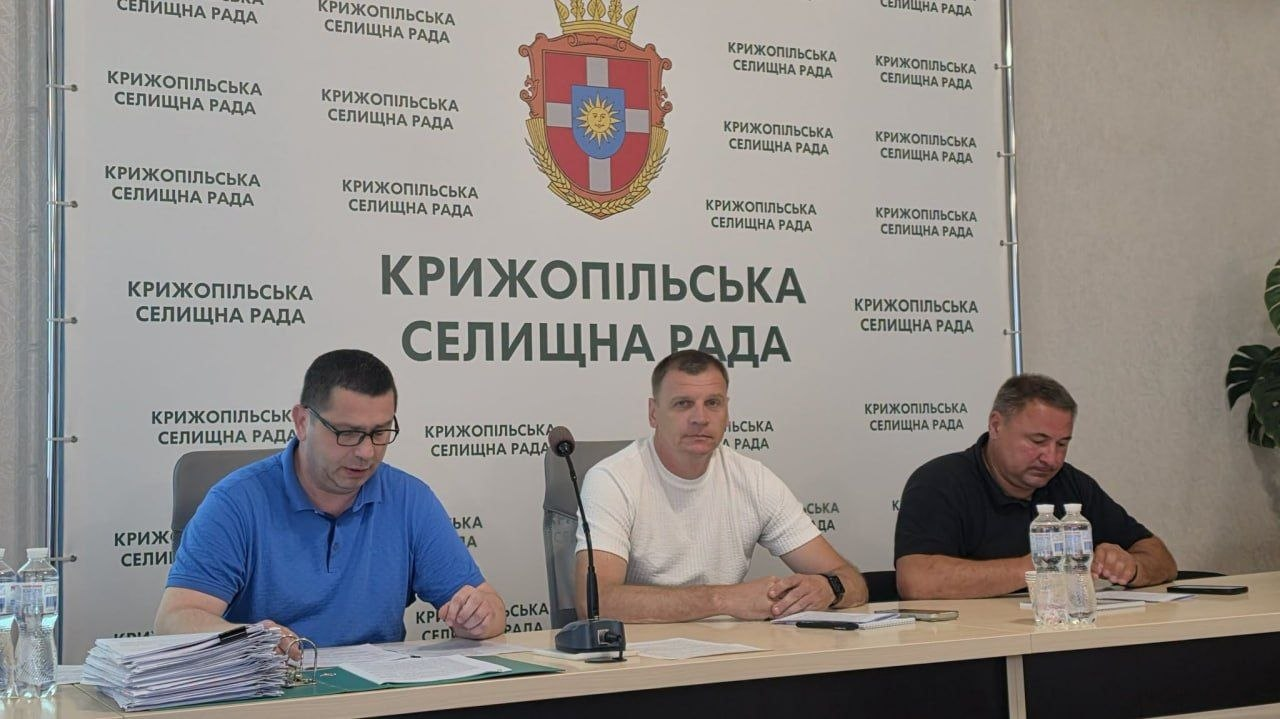 Три чоловіки сидять за столом під час робочого засідання на тлі брендованого банера з гербом громади. Вони працюють із документами, перед ними встановлено мікрофон та стоїть вода.