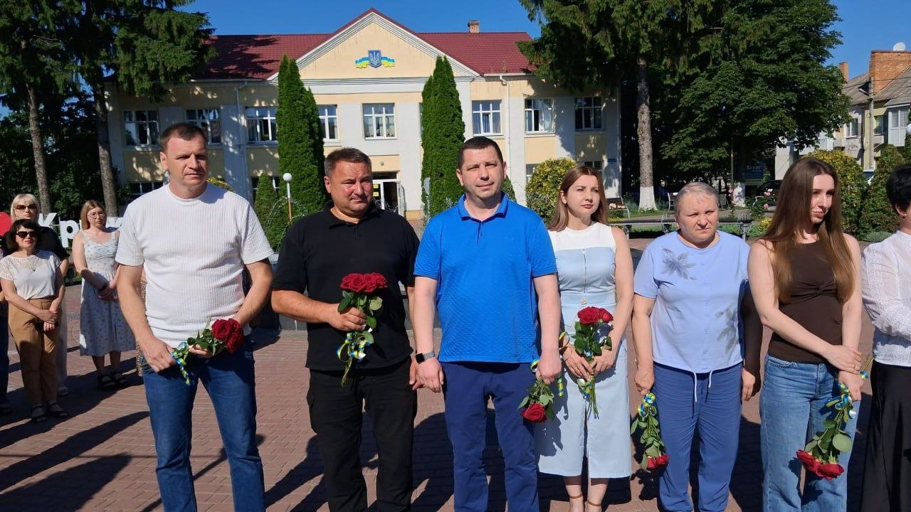 Група людей стоїть на площі просто неба перед двоповерховою адміністративною будівлею з гербом України на фасаді. Кілька осіб у першому ряду тримають у руках червоні троянди, перев'язані синьо-жовтими стрічками. Подія відбувається у сонячний день на тлі зелених дерев.