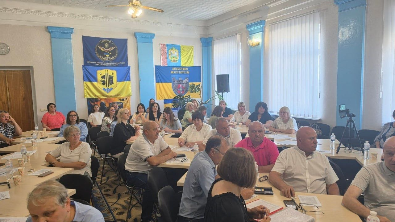 Велика група людей сидить за робочими столами під час засідання у залі з блакитними колонами. Присутні слухають доповідача, дехто робить записи у документах. На задньому плані ліворуч розвішані військові та державні прапори, а біля вікон праворуч встановлено штатив із телефоном.
