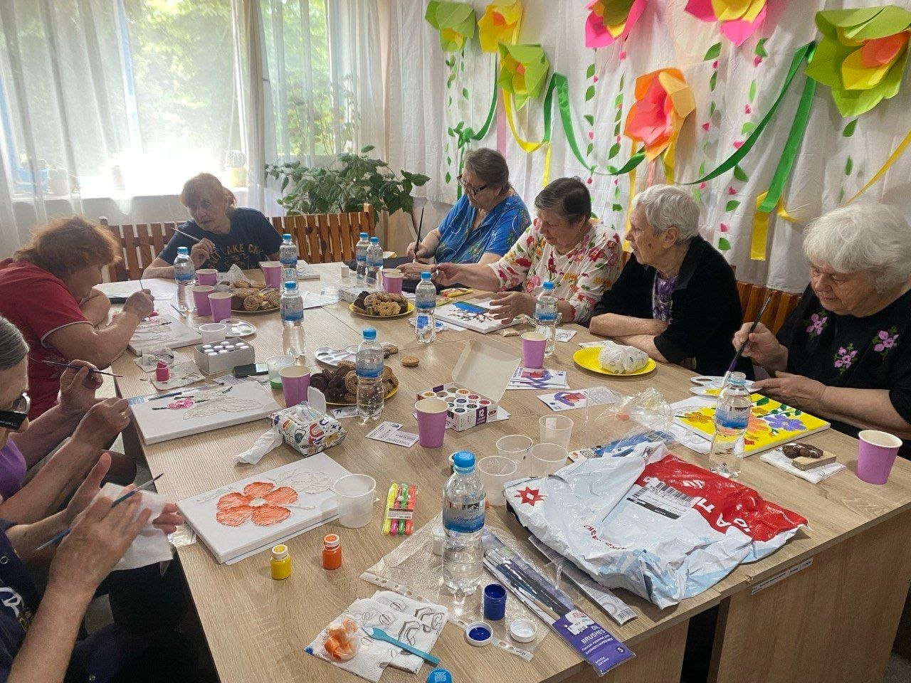 Група літніх жінок сидить за великим столом під час творчого заняття з малювання. Вони тримають пензлі та розфарбовують невеликі малюнки на папері й полотнах. На столі розкладені художні матеріали, пляшки з водою та солодощі, а стіна на задньому плані прикрашена кольоровими паперовими квітами.