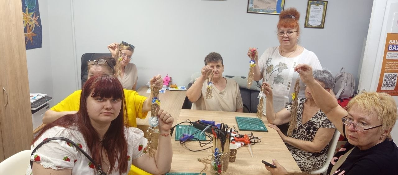 Група жінок за столом у кабінеті демонструє виготовлені власноруч ляльки-обереги з ниток. На столі перед ними лежать інструменти для рукоділля, зокрема ножиці та клейовий пістолет.