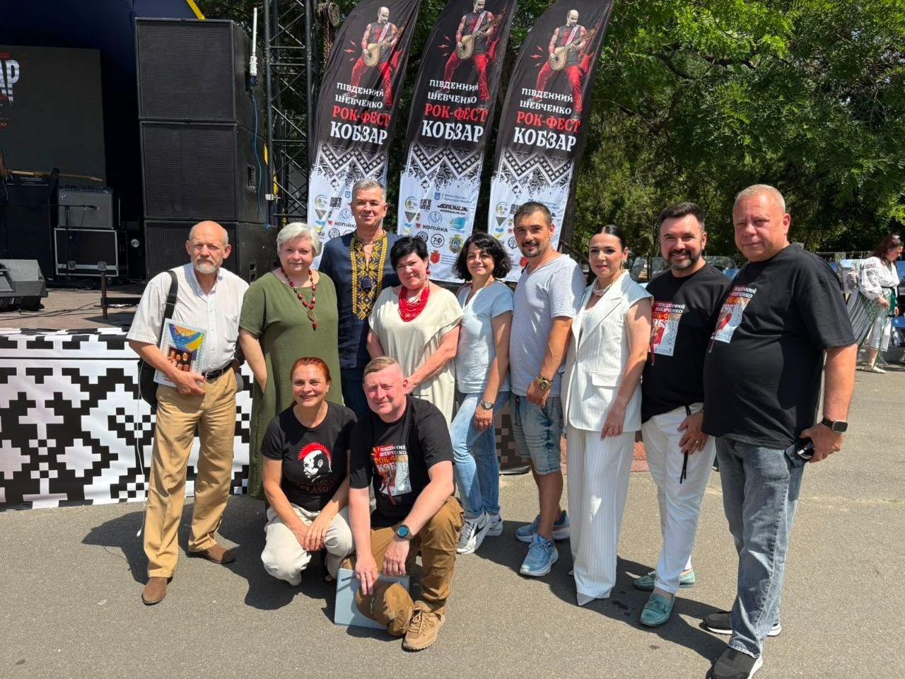 Група людей позирує для спільного фото просто неба біля фестивальної сцени. Позаду них видно вертикальні банери з написом «Південний Шевченко Рок-Фест Кобзар». Подія відбувається у сонячний день на асфальтованому майданчику.