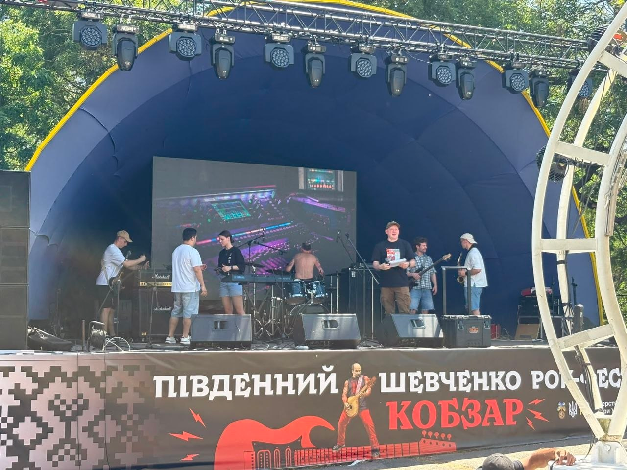 Музичний гурт готується до виступу або проводить репетицію на великій критій сцені просто неба. На сцені встановлено звукову та світлову апаратуру, великий екран, а попереду розміщено банер із написом «Південний Шевченко Рок-Фест Кобзар». Подія відбувається у сонячний день.