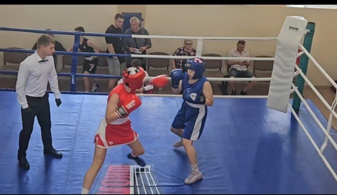 Два боксери у захисних шоломах, червоній та синій формах, проводять поєдинок на синьому ринзі. Зліва за ними спостерігає рефері у білій сорочці та чорних штанах, а на задньому плані за канатами сидять глядачі.