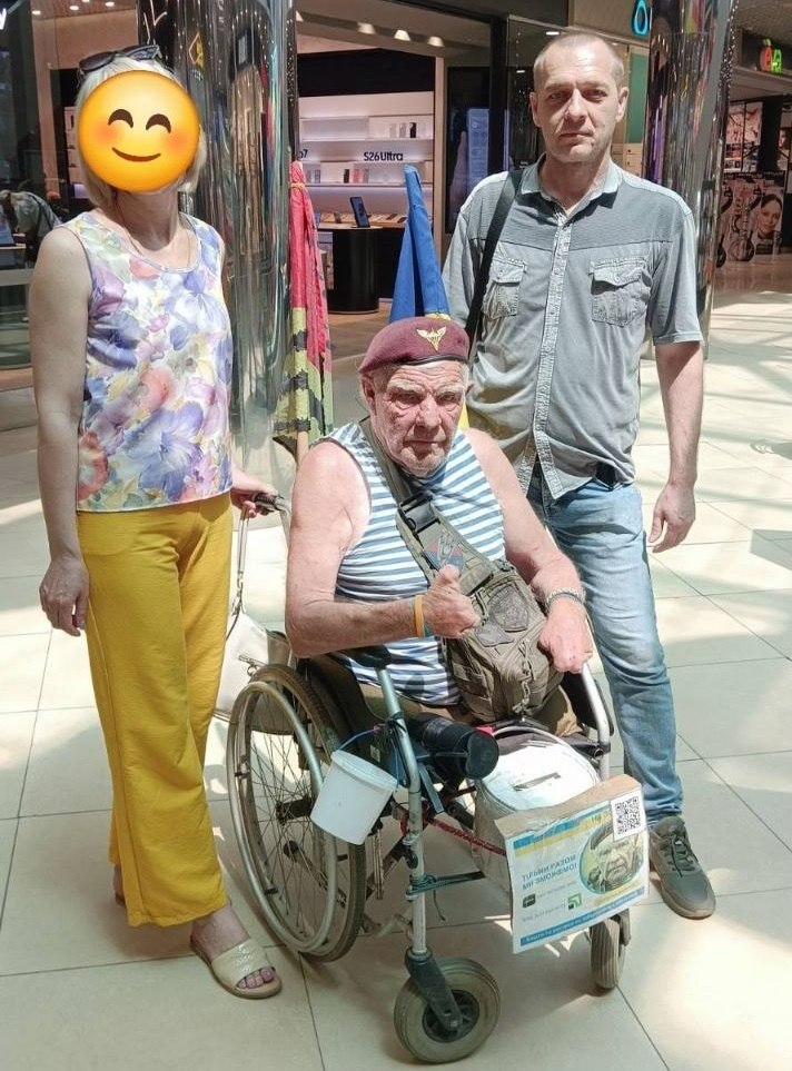 Літній чоловік у смугастій майці, бордовому береті та у кріслі колісному сидить у ТРЦ, показуючи великий палець угору. Поруч із ним стоять жінка у жовтих штанах (обличчя приховане емодзі) та чоловік у сірій сорочці й джинсах. На кріслі колісному закріплено невелику табличку зі збором коштів, а на задньому плані видно прапори та вітрини магазинів.