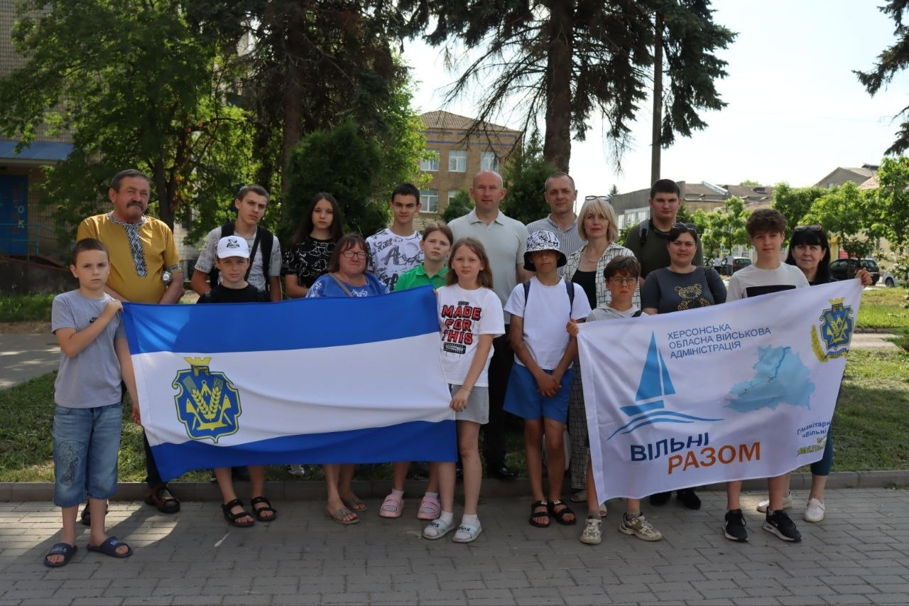 Група дорослих та дітей стоїть на вулиці біля дерев і тримає перед собою два прапори. Ліворуч розгорнуто синьо-білий прапор із гербом Херсонської області, а праворуч — білий прапор із написом «Херсонська обласна військова адміністрація», силуетом вітрильника та гаслом «Вільні разом». Усі присутні вбрані у літній повсякденний одяг.
