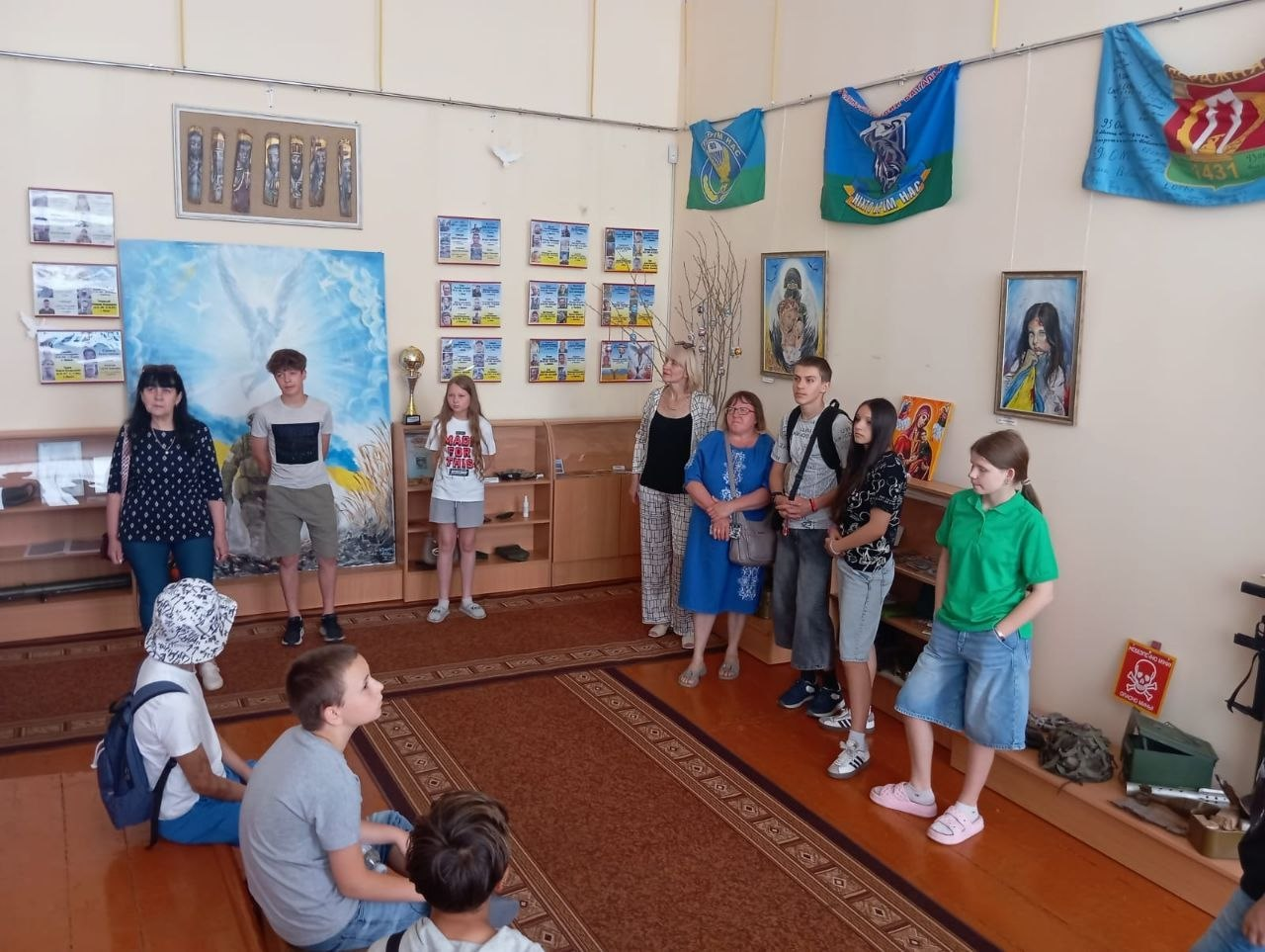 Група дітей та дорослих перебуває у музейній кімнаті під час екскурсії. Частина присутніх стоїть уздовж стін, а троє дітей сидять на килимі по центру. На стінах приміщення розміщені картини, плакати, фотографії у рамках та військові прапори під стелею.