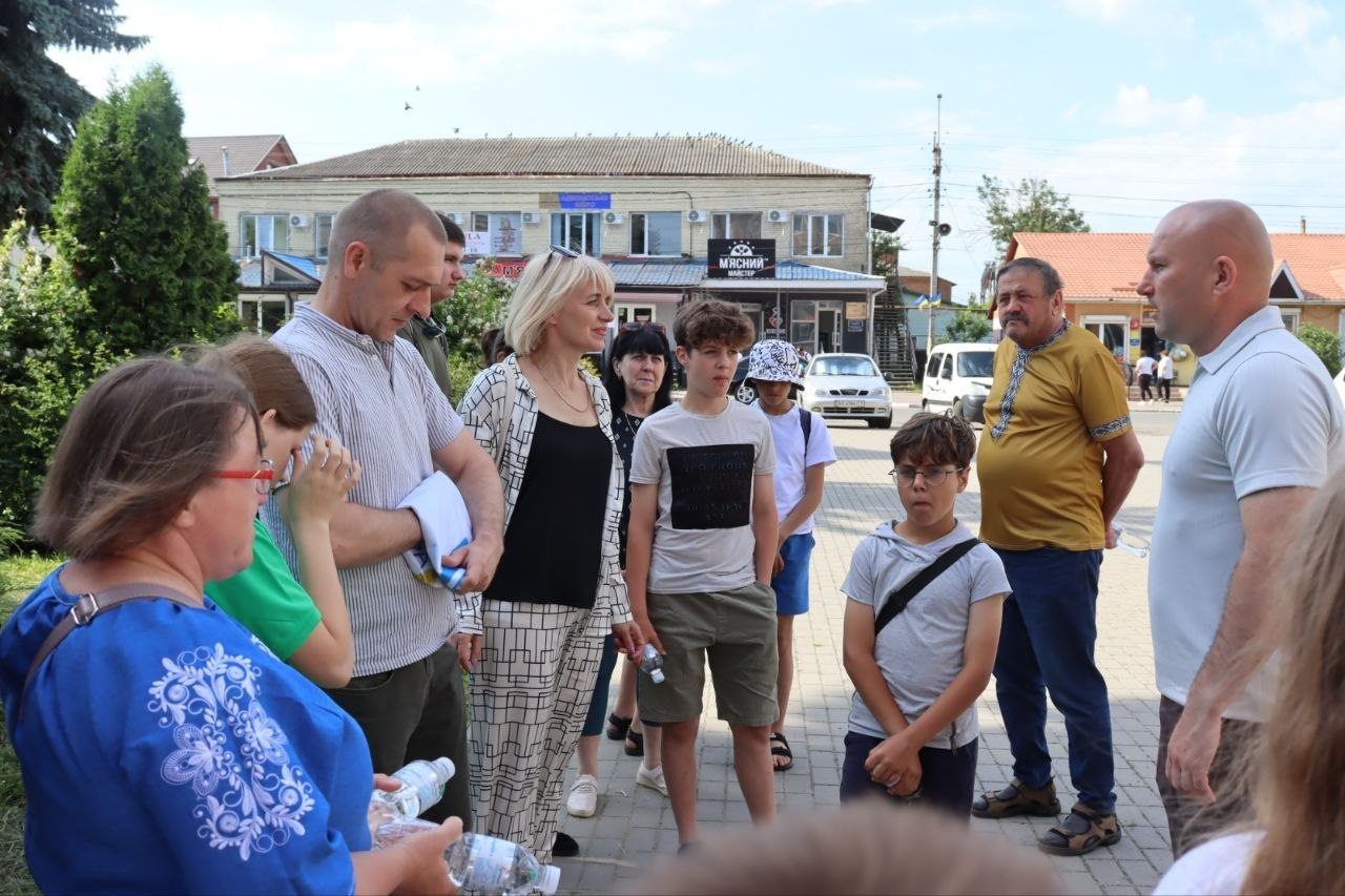 Група дорослих та дітей стоїть на вулиці та спілкується на тлі будівель, дерев та автомобілів у сонячний день.