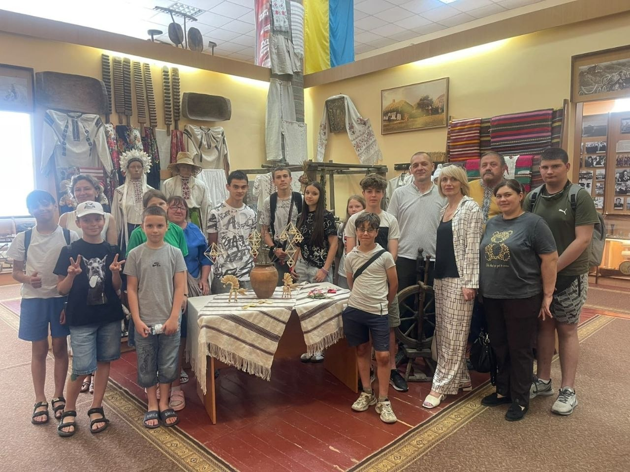 Велика група людей різного віку стоїть разом для спільного фото в залі музею на тлі українського прапора та етнографічних експонатів.