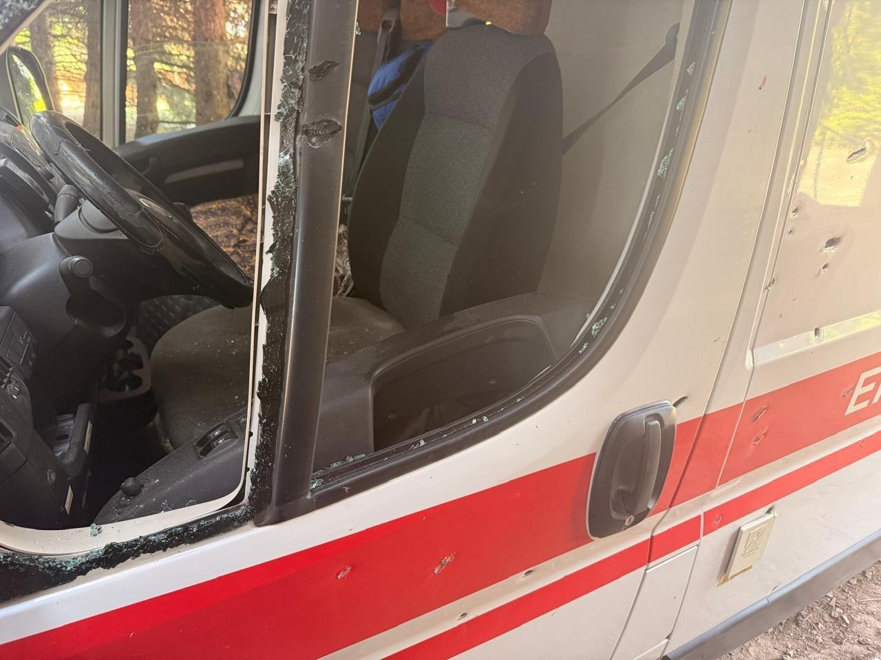 Пошкоджений білий автомобіль швидкої допомоги з червоною смугою, розбитим боковим склом та слідами від уламків або куль на дверцятах.