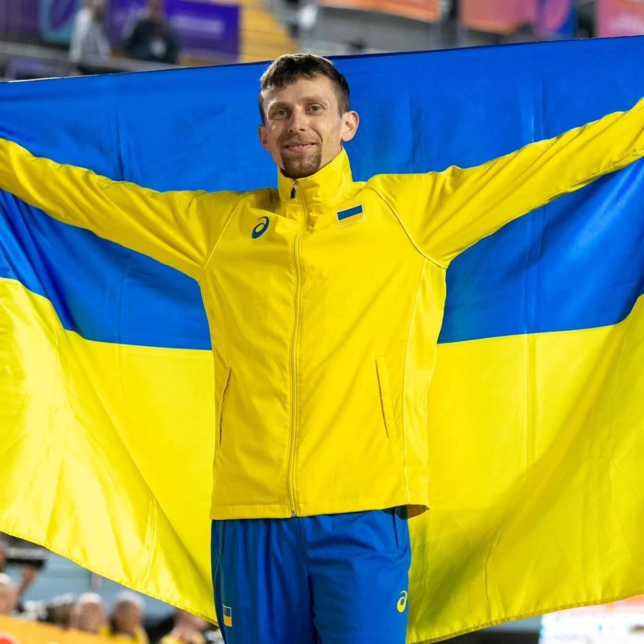 Спортсмен у синьо-жовтій спортивній формі посміхається та тримає за спиною великий прапор України.