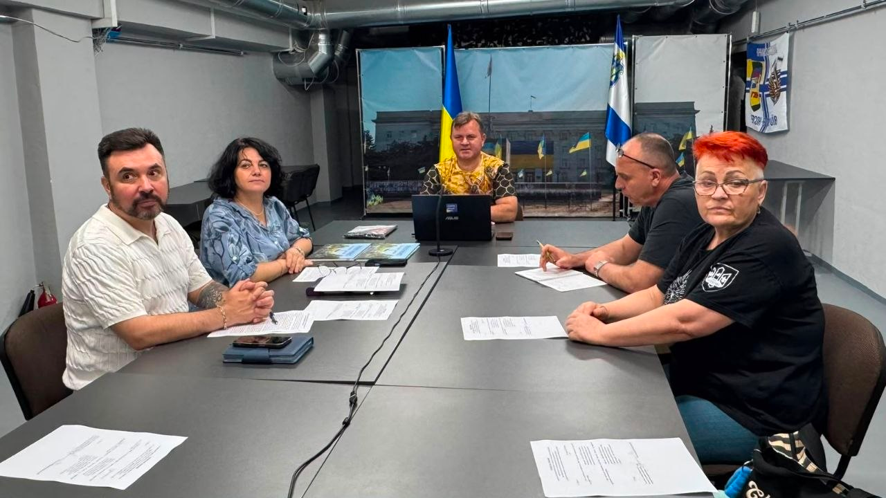 Група людей сидить за великим Т-подібним столом із документами під час наради на тлі прапорів та зображення будівлі.