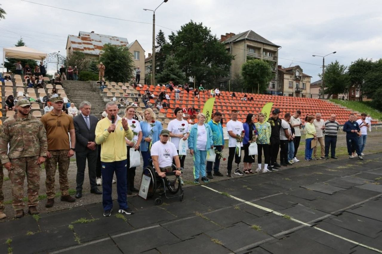 Офіційне відкриття заходу на стадіоні, де перед трибунами в ряд стоять люди, серед яких військові, цивільні та чоловік у кріслі колісному, а один із присутніх говорить у мікрофон.