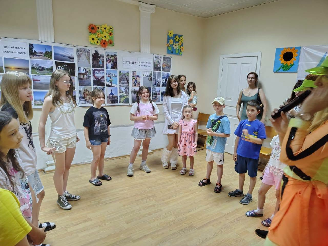 Група дітей та дорослих стоїть півколом у кімнаті під час заходу, де ведуча в яскравому костюмі говорить у мікрофон, а на стіні позаду розміщено стенди з фотографіями.
