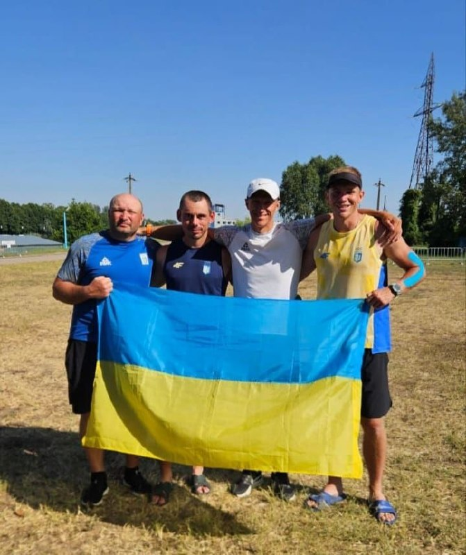 Чотири чоловіки у спортивному одязі стоять разом на відкритому полі в сонячний день і тримають перед собою великий прапор України.