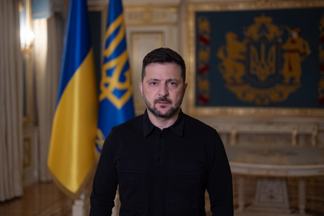 Чоловік у чорній сорочці стоїть у приміщенні на тлі державного прапора України та штандарта, а на стіні позаду видно велике зображення герба.