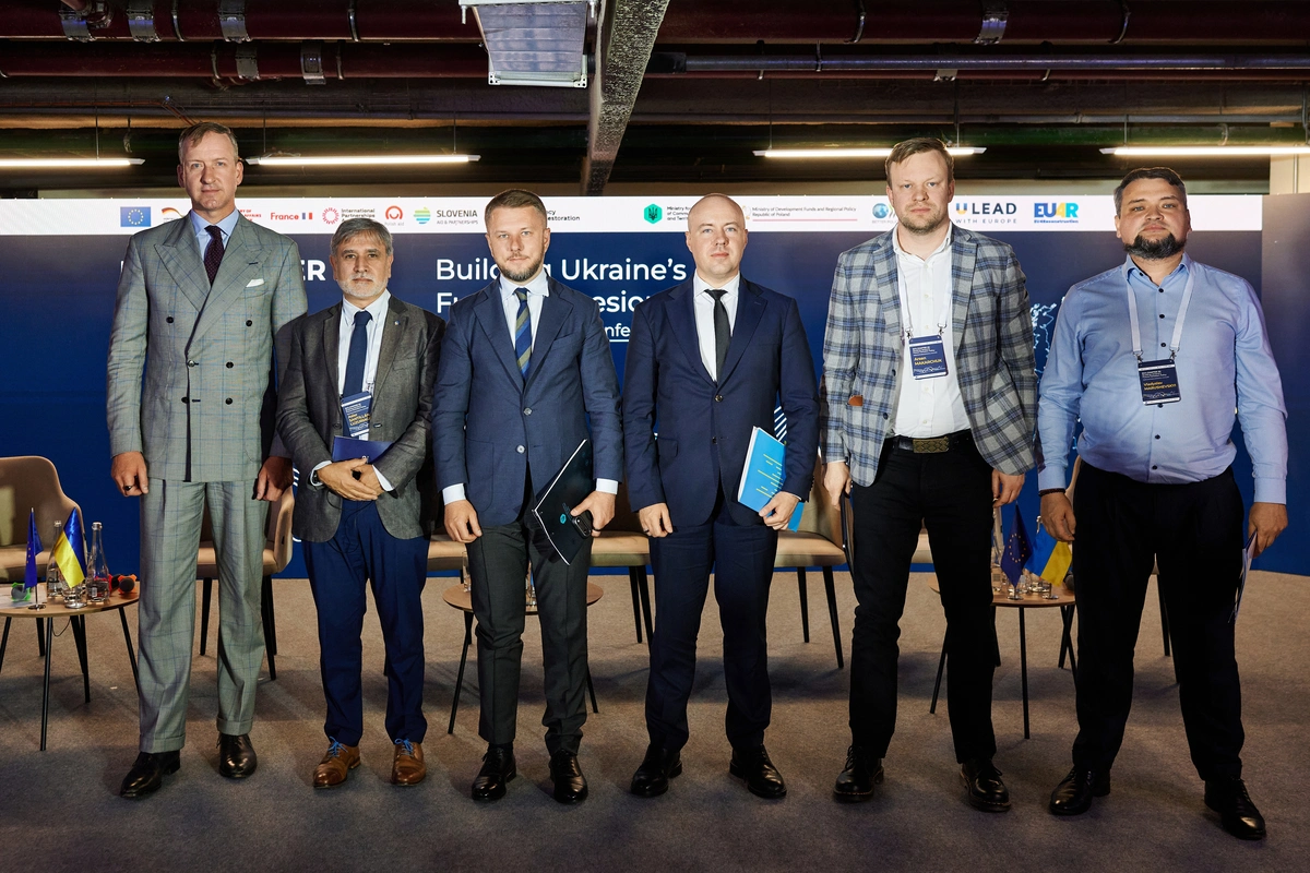 Шість чоловіків у ділових костюмах стоять у ряд на сцені конференції на тлі великого синього банера з логотипами та англомовним текстом.