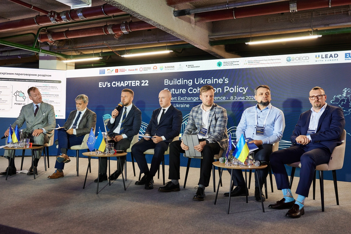 Сім спікерів у діловому одязі сидять на сцені конференції на тлі великого синього банера з написами «EU's CHAPTER 22» та «Building Ukraine's Future Cohesion Policy».