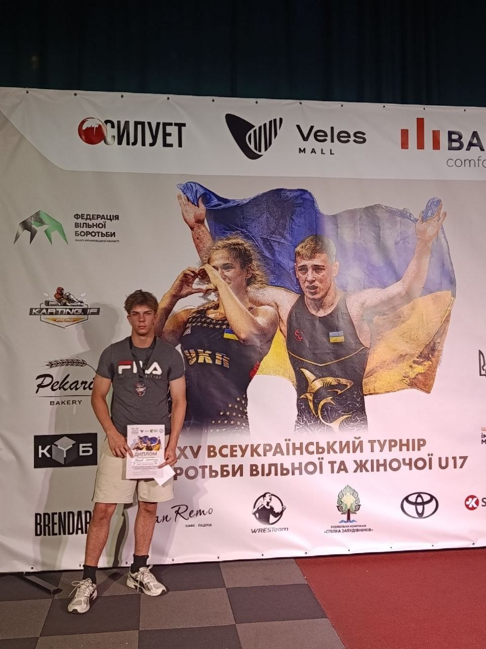 Молодий спортсмен у сірій футболці та шортах стоїть перед великим банером спортивного турніру, тримаючи в руках диплом. На банері позаду нього зображені борці з українським прапором та розміщені логотипи спонсорів.