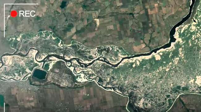 Супутниковий знімок обмілілого русла річки Дніпро та колишнього Каховського водосховища з оголеним дном, де звивається тонка смуга води. Навколо видно прилеглі зелені зони, сільськогосподарські поля та населені пункти, а у верхньому лівому кутку розміщено червоний значок «REC».