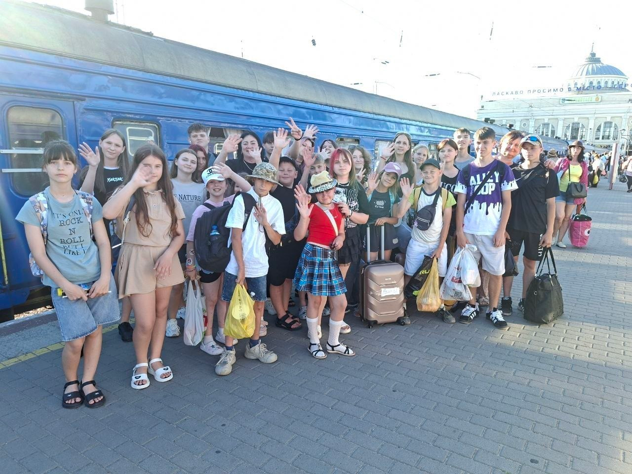 Велике скупчення дітей та підлітків із сумками й пакетами стоїть на залізничному пероні поруч із синім пасажирським вагоном. Більшість із них посміхаються та махають руками в камеру, а на задньому плані видніється будівля вокзалу.