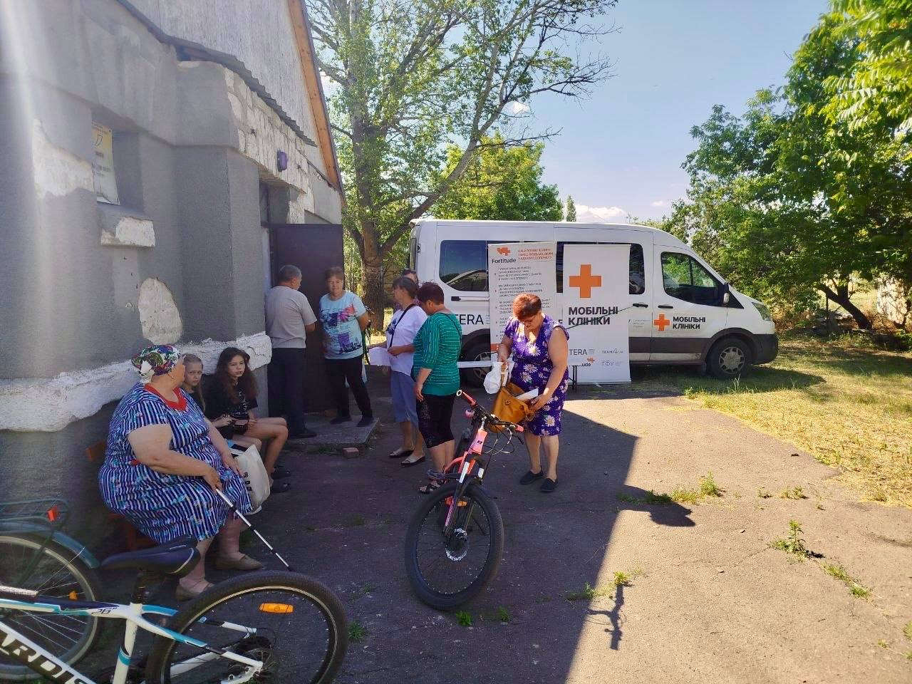 Група місцевих жителів зібралася біля будівлі та білого мікроавтобуса з написом «Мобільні клініки» та червоним хрестом. Люди спілкуються між собою, дехто чекає на лавці або стоїть біля входу, а на передньому плані видно велосипеди на тлі зелених дерев у сонячний день.