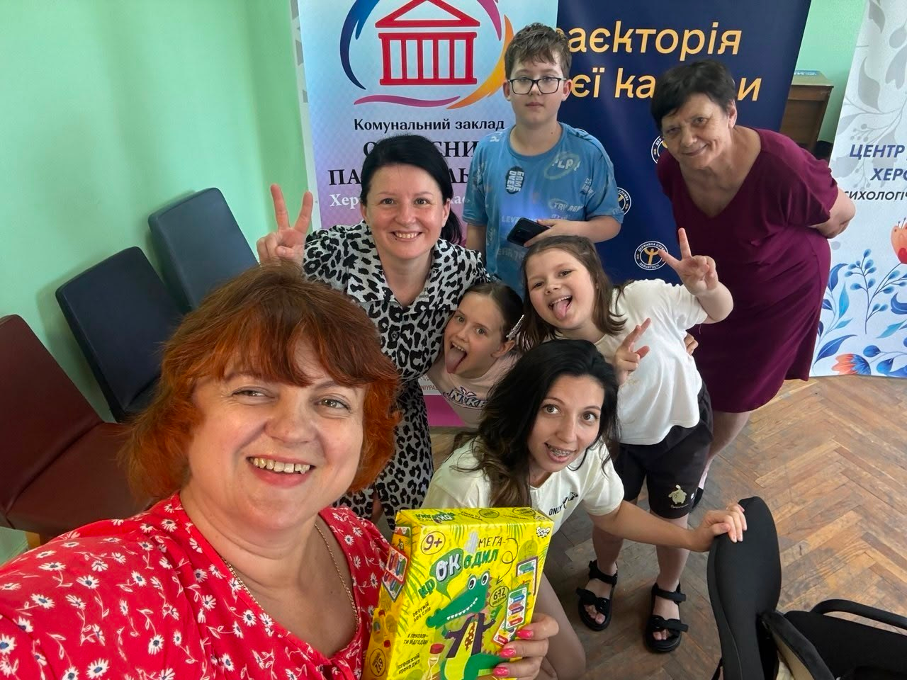 Група дітей та дорослих позирує для спільного селфі у світлій кімнаті. Жінка на передньому плані тримає жовту коробку з настільною грою «Крокодил», а інші учасники на фоні посміхаються та показують жест «мир» (V). На задньому плані видно офісні стільці та великі інформаційні банери з написами українською мовою.