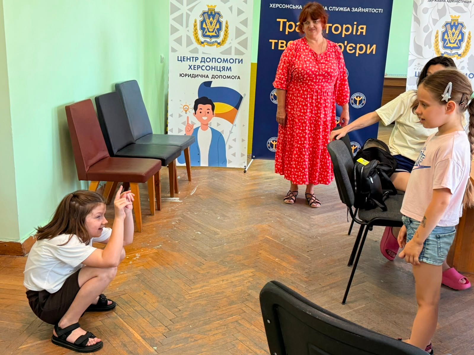 Група дітей та дорослих у світлій кімнаті під час заходу в гуманітарному центрі. Ліворуч на підлозі присіла дівчинка, яка показує загадане слово жестами, а праворуч стоїть інша дитина та спостерігає. На задньому плані стоїть жінка у червоній сукні, розміщені офісні стільці та інформаційні банери з написами «Центр допомоги херсонцям» та «Траєкторія твого успіху».