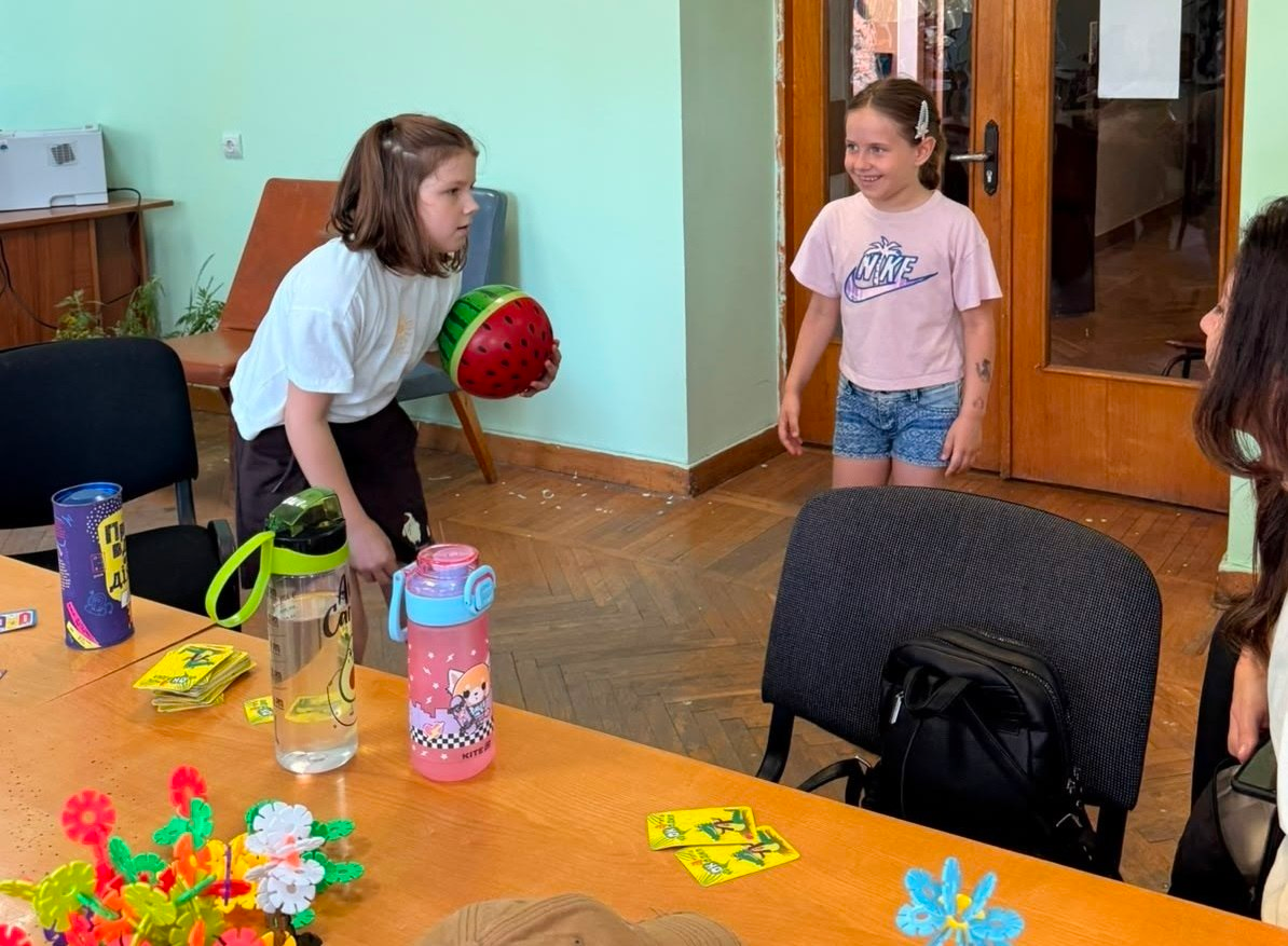 Дві дівчинки грають у рухливу гру у світлій кімнаті. Ліворуч дитина тримає в руках м'яч, розфарбований під кавун, а інша дівчинка праворуч посміхається та спостерігає за нею. На передньому плані видно стіл, на якому стоять пляшки для води, конструктор та розкладені картки для настільної гри.