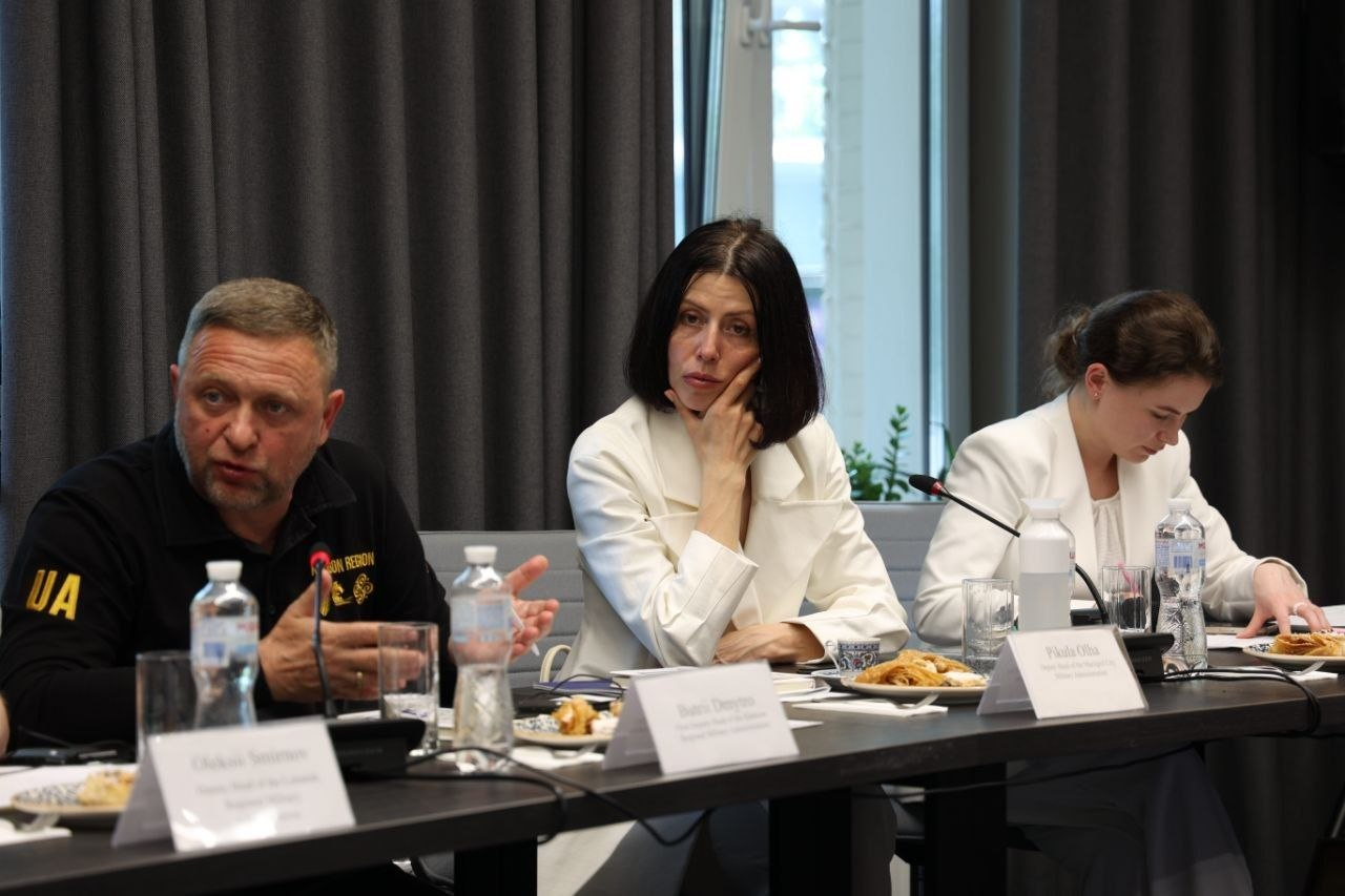 Три особи сидять за столом під час засідання. Чоловік ліворуч у чорному одязі виступає перед мікрофоном, а дві жінки у світлих костюмах поруч уважно слухають. На столі перед ними стоять пляшки з водою, тарілки з їжею та іменні таблички.