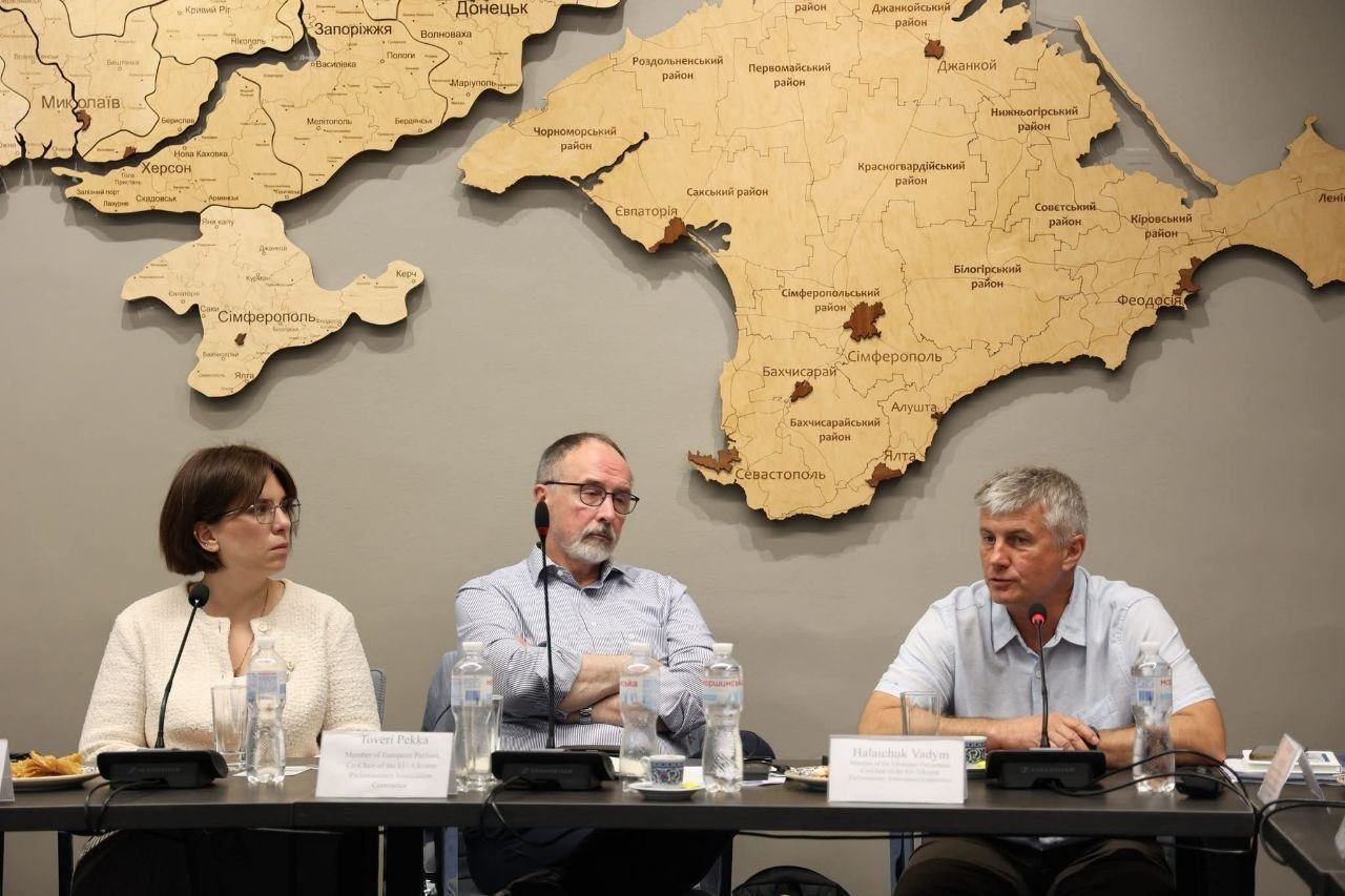 Три особи сидять за круглим або довгим столом під час конференції чи офіційного засідання. На передньому плані перед ними встановлено мікрофони, пляшки з водою та розставлено іменні таблички. На сірій стіні за їхніми спинами розміщена велика дерев'яна мапа України, де детально видно Кримський півострів та південні області.