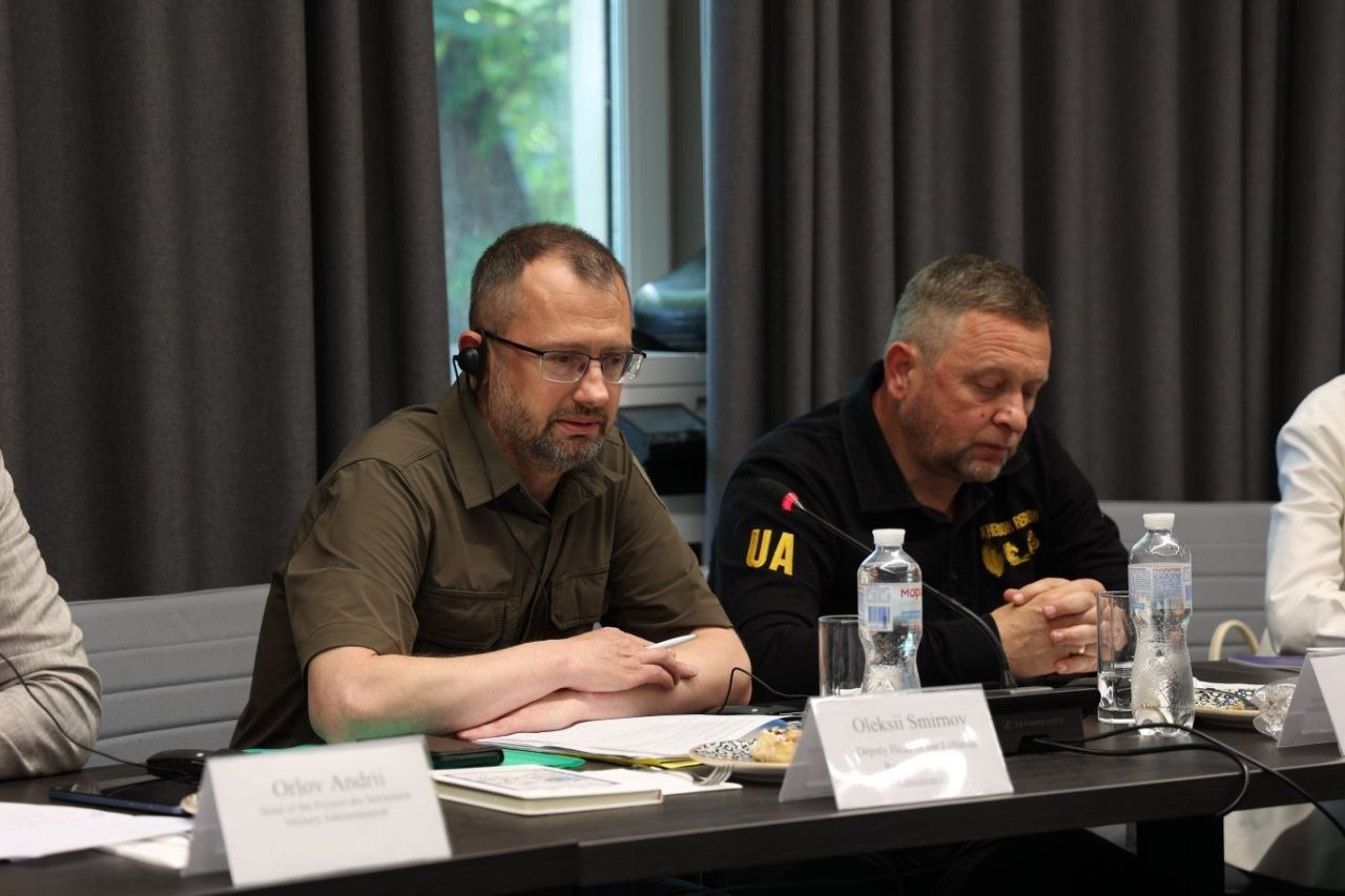 Два чоловіки сидять за робочим столом під час засідання чи офіційної зустрічі. Чоловік ліворуч у зеленій сорочці мілітарі та окулярах тримає ручку і дивиться на документи, а чоловік праворуч у чорному одязі з написами уважно слухає. На столі перед ними стоять мікрофони, пляшки з водою, склянки та іменні таблички.
