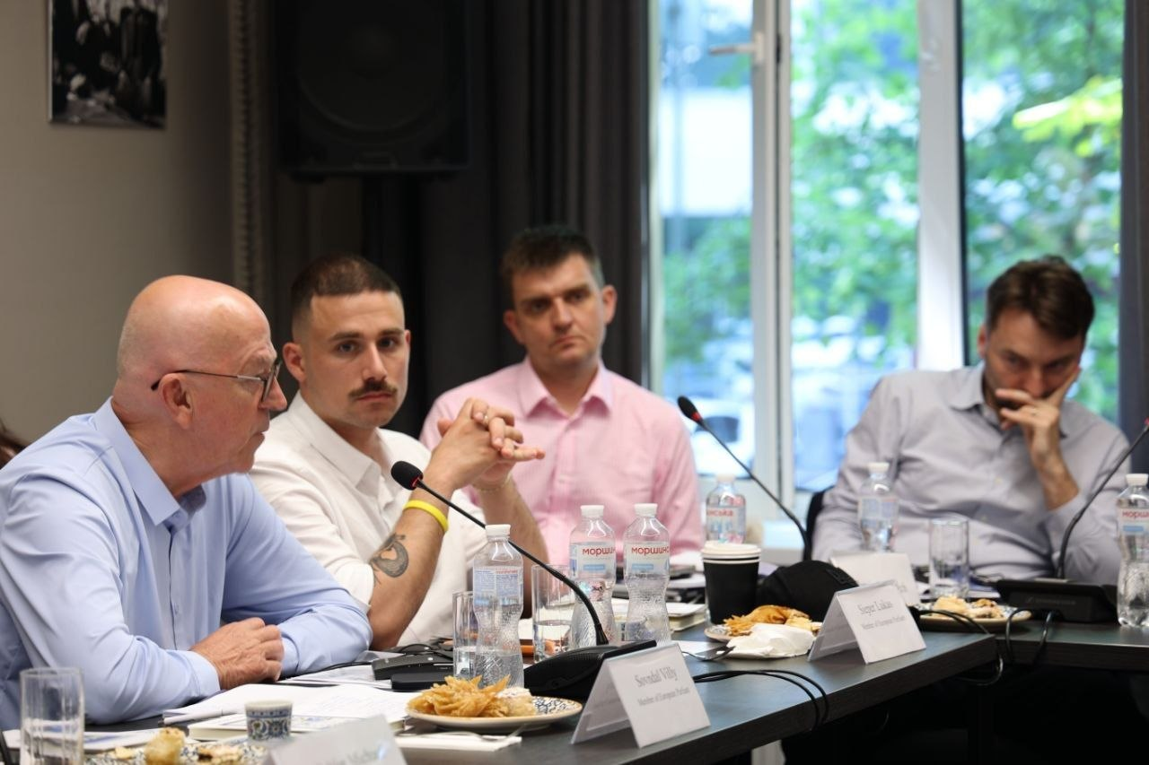 Чотири чоловіки в сорочках сидять за столом під час ділової зустрічі чи конференції в залі з великим вікном. На передньому плані ліворуч старший чоловік у блакитній сорочці висловлює свою думку, тоді як інші учасники зосереджено слухають його. На столі перед ними розставлені мікрофони, пляшки з водою, іменні таблички та тарілки з їжею.