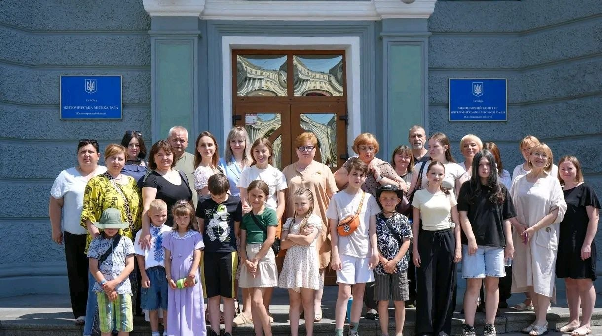 Групове фото великої компанії дорослих та дітей різного віку на сходах перед головним входом до офіційної установи. На світло-синій стіні будівлі по обидва боки від дерев'яних дверей висять сині інформаційні таблички з гербом України. Люди одягнені у літній повсякденний одяг, дехто з дітей має головні убори, а одна дівчинка попереду вбрана у довгу фіолетову сукню.