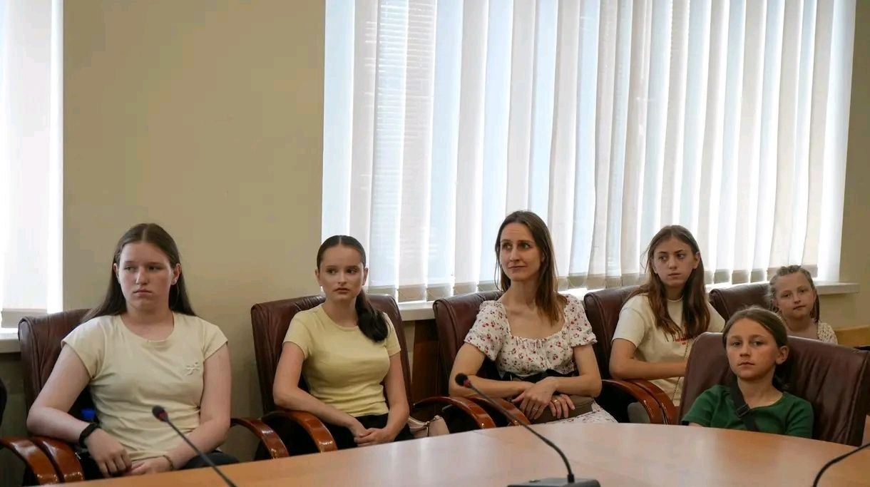 Жінка та п’ять дівчат різного віку сидять у ряд у коричневих шкіряних кріслах за великим дерев'яним столом для нарад, на якому встановлені мікрофони. Вони вдягнені у літній повсякденний одяг світлих та зелених відтінків. На задньому плані видно світлу стіну та велике вікно, завішене білими вертикальними жалюзі.
