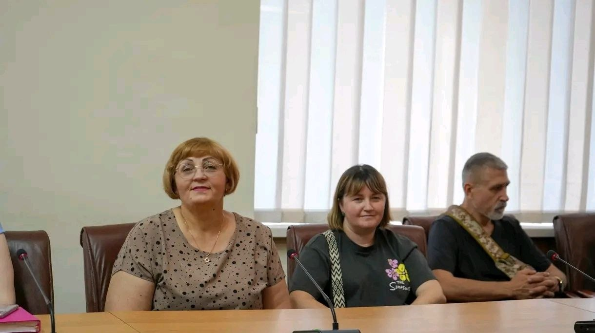 Дві жінки та чоловік сидять за столом для нарад із мікрофонами на тлі вікна з білими вертикальними жалюзі. Одна з жінок в окулярах та блузі в цятку, інша — у темній футболці, а чоловік — у чорній сорочці.