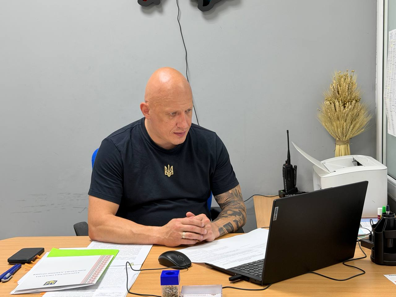 Чоловік у чорній футболці з гербом України сидить за робочим столом, дивиться на екран ноутбука та тримає руки зчепленими перед собою. На столі лежать документи, комп'ютерна миша, рація та канцелярське приладдя, а на задньому плані видно принтер і декоративний букет з колосків.