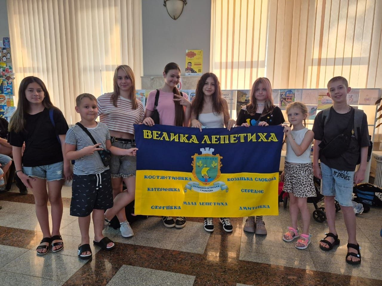 Група дітей та підлітків стоїть у приміщенні й тримає синьо-жовтий прапор із написом «Велика Лепетиха» та гербом. На прапорі також перелічені назви інших населених пунктів громади. Діти в літньому одязі посміхаються та позирують для фото на тлі вікон і дитячих малюнків.