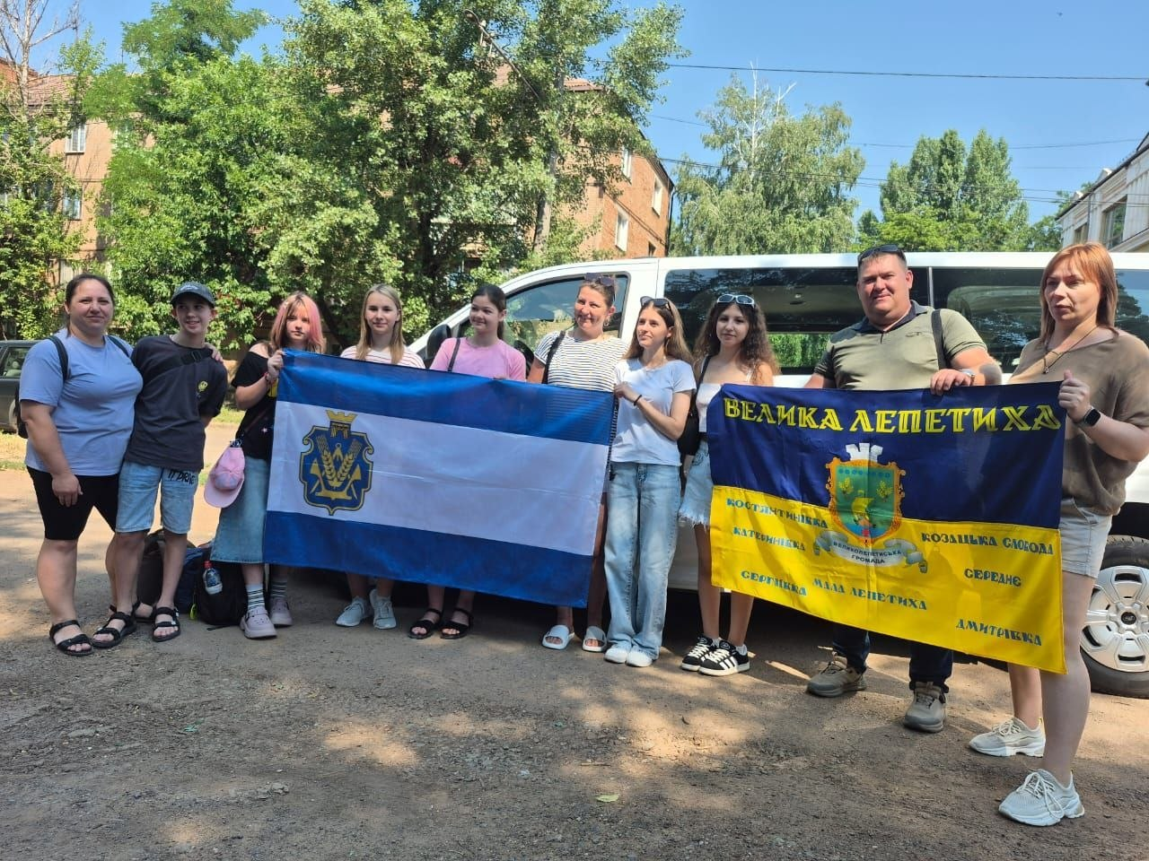Група дорослих та підлітків у літньому одязі стоїть на вулиці біля білого мікроавтобуса, тримаючи два прапори: біло-синій із гербом та синьо-жовтий із написом «Велика Лепетиха».