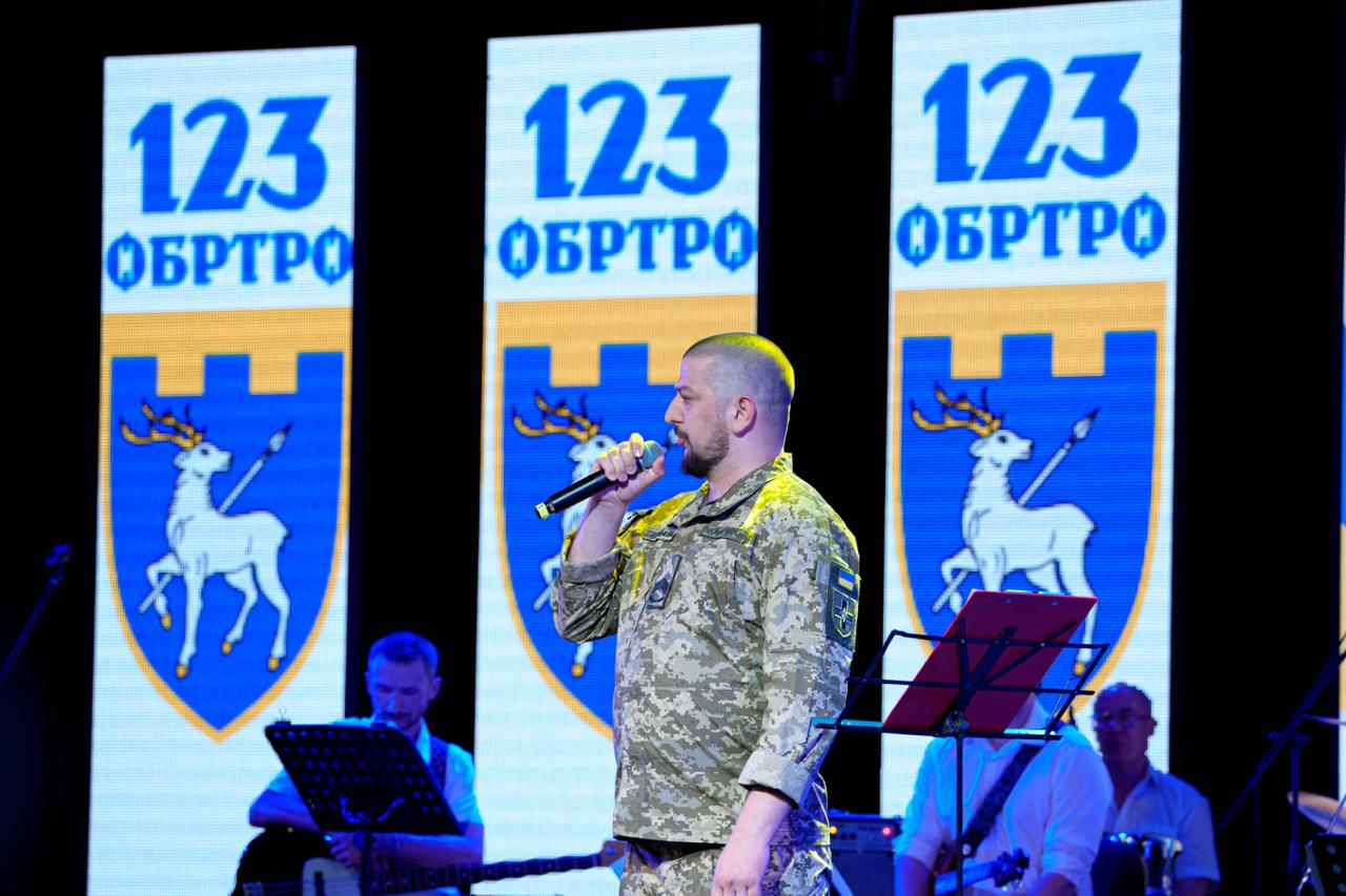 Чоловік у військовій формі виступає на сцені з мікрофоном у руці на тлі трьох великих екранів із символікою «123 ОБРТрО» та гербом. На задньому плані з обох боків від нього видно музикантів із гітарами та пюпітрами.