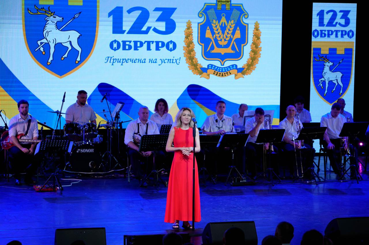 Жінка в довгій червоній сукні співає на сцені перед мікрофоном на тлі великого екрана з написом «123 ОБРТрО Приречена на успіх!», гербом Херсонської області та символікою бригади. За нею на сцені розмістився музичний оркестр з інструментами та пюпітрами.