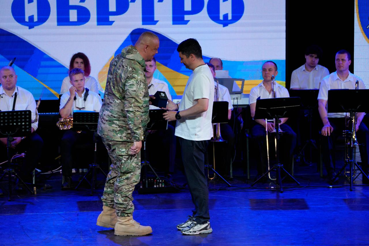 Чоловік у камуфляжній формі та берцах передає нагороду чоловікові в білій футболці та темних штанах на сцені. На задньому плані сидить музичний оркестр у білих сорочках перед великим екраном із синьо-жовтими елементами.