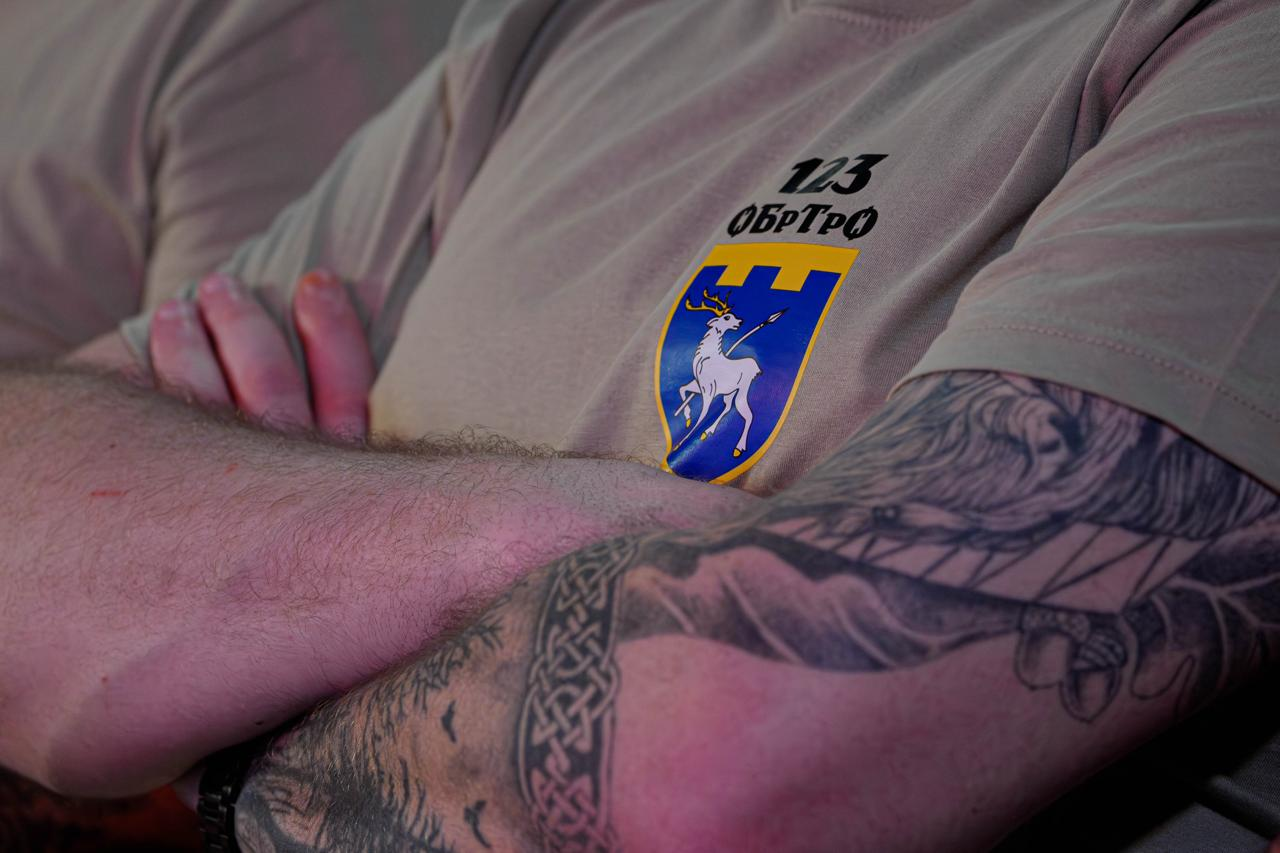 Крупний план чоловіка зі схрещеними на грудях руками, на одній з яких видно велике татуювання. Він одягнений у бежеву футболку з логотипом «123 ОБрТрО», що містить напис та герб із зображенням білого оленя на синьому щиті.