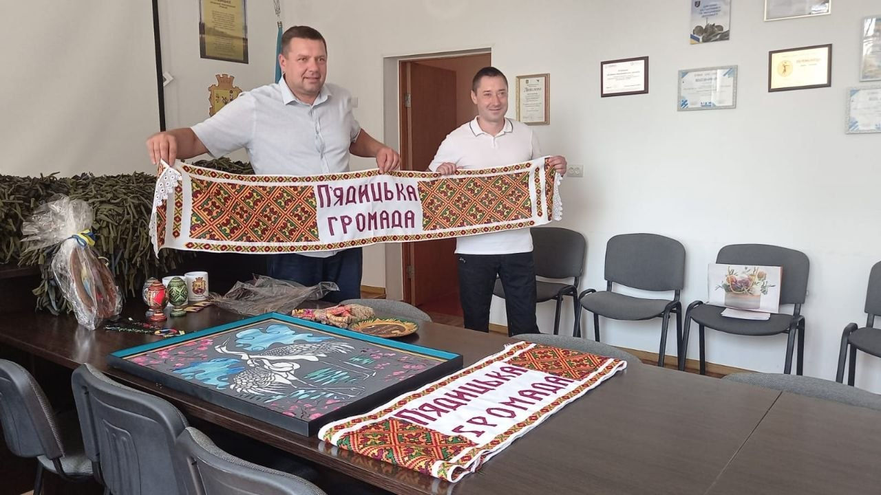 Два чоловіки у кабінеті тримають вишитий рушник із написом «П'ядицька громада». На столі перед ними лежать українські сувеніри: ще один аналогічний рушник, картина із зображенням лелек, розписані писанки та інші вироби традиційного мистецтва.