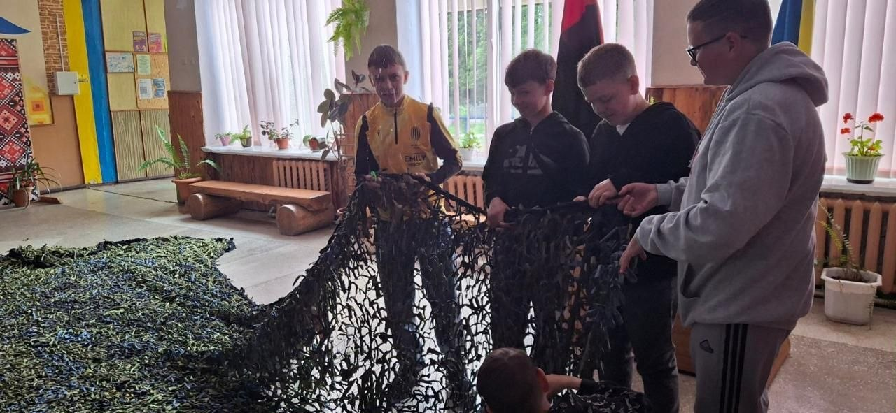 Четверо хлопців у приміщенні разом тримають та розправляють велику маскувальну сітку зеленого та чорного кольорів. Частина сітки розстелена на підлозі, а на задньому плані видно кімнатні рослини та патріотичний декор.