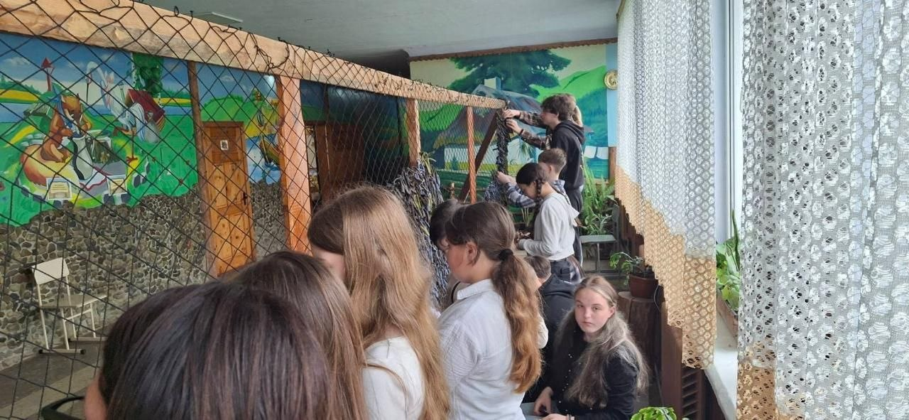 Група дітей та підлітків у коридорі приміщення разом плетуть велику маскувальну сітку, закріплену на дерев'яному каркасі. На стіні позаду них зображено яскравий мурал у традиційному українському стилі з пейзажами та персонажами казок.